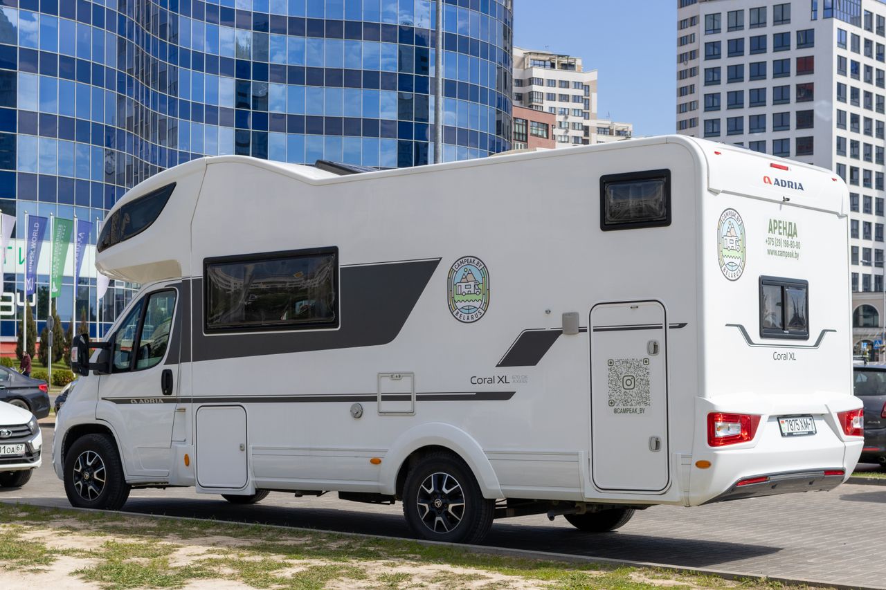A white RV parked beside modern glass buildings in a sunny urban setting.