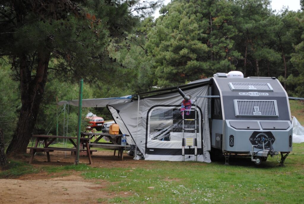 A caravan set up in a tranquil forest campsite with wooden tables and lush greenery.